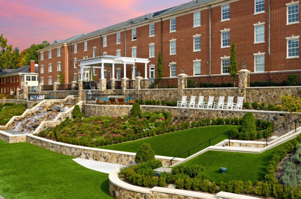 The McCallie School in Chattanooga, TN is a three-story brick building that sits along The Keith Greenway. There are stone walls, lush green grass, and several rocking chairs in front of the school.